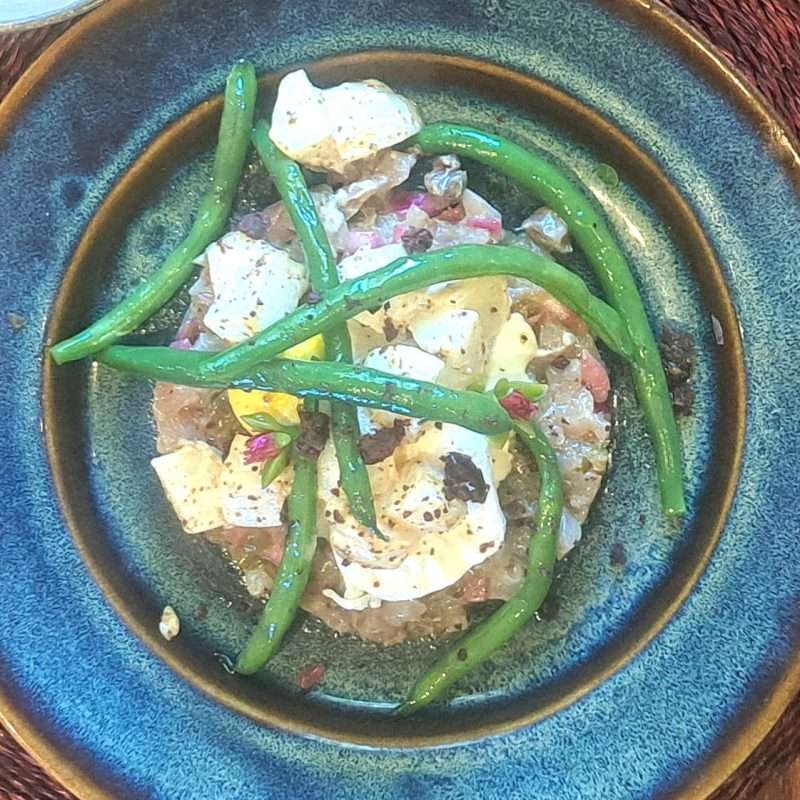 A beautifully plated dish of cured yellowtail with pickled red onion, capers, poached egg, potato, and green beans, served on a deep blue ceramic plate.