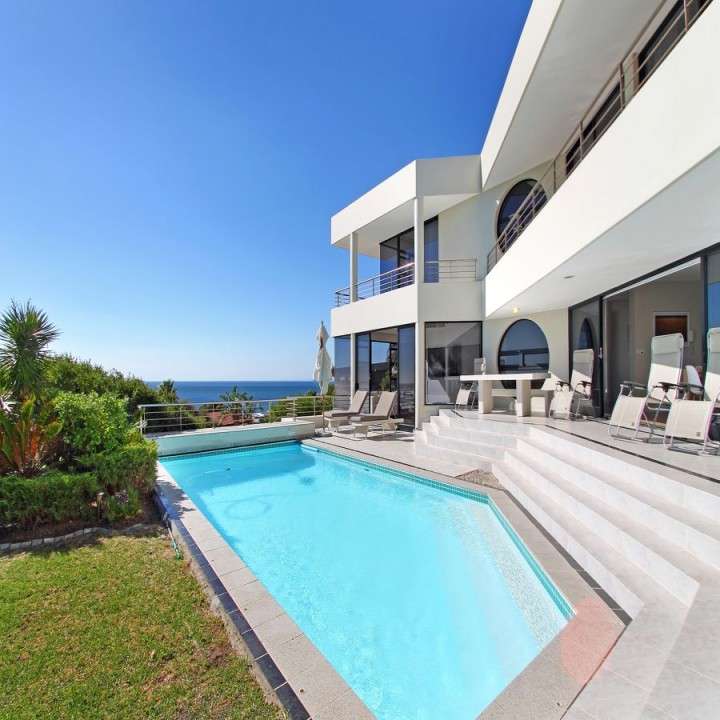 A luxurious modern villa with a sleek white exterior, large glass windows, and an inviting swimming pool overlooking the ocean. The outdoor space features sun loungers, a dining area, and lush greenery under a bright blue sky